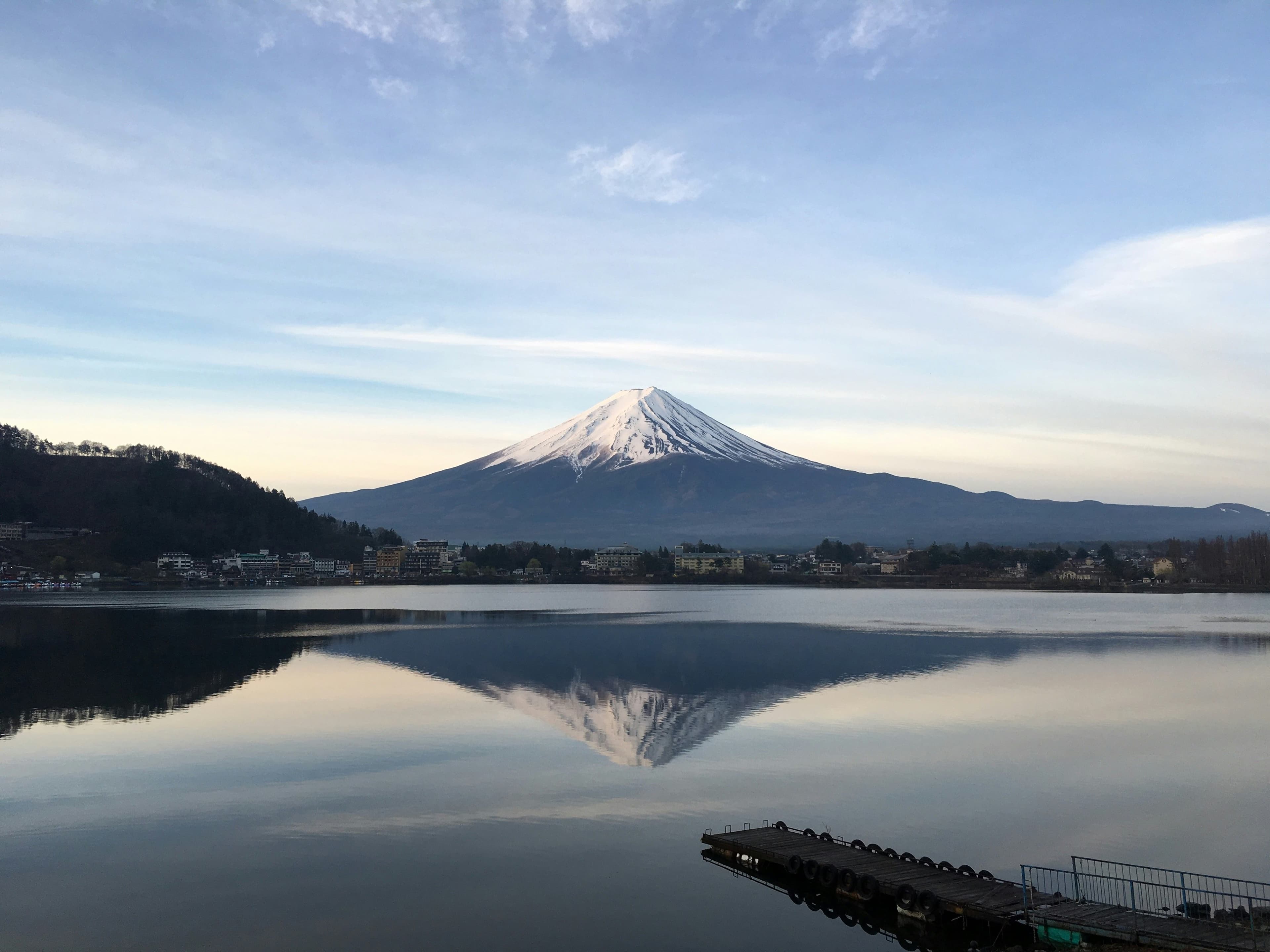 El Monte Fuji nevado reflejado en el agua tranquila del lago Kawaguchiko al amanecer, con un pequeño muelle de madera en primer plano