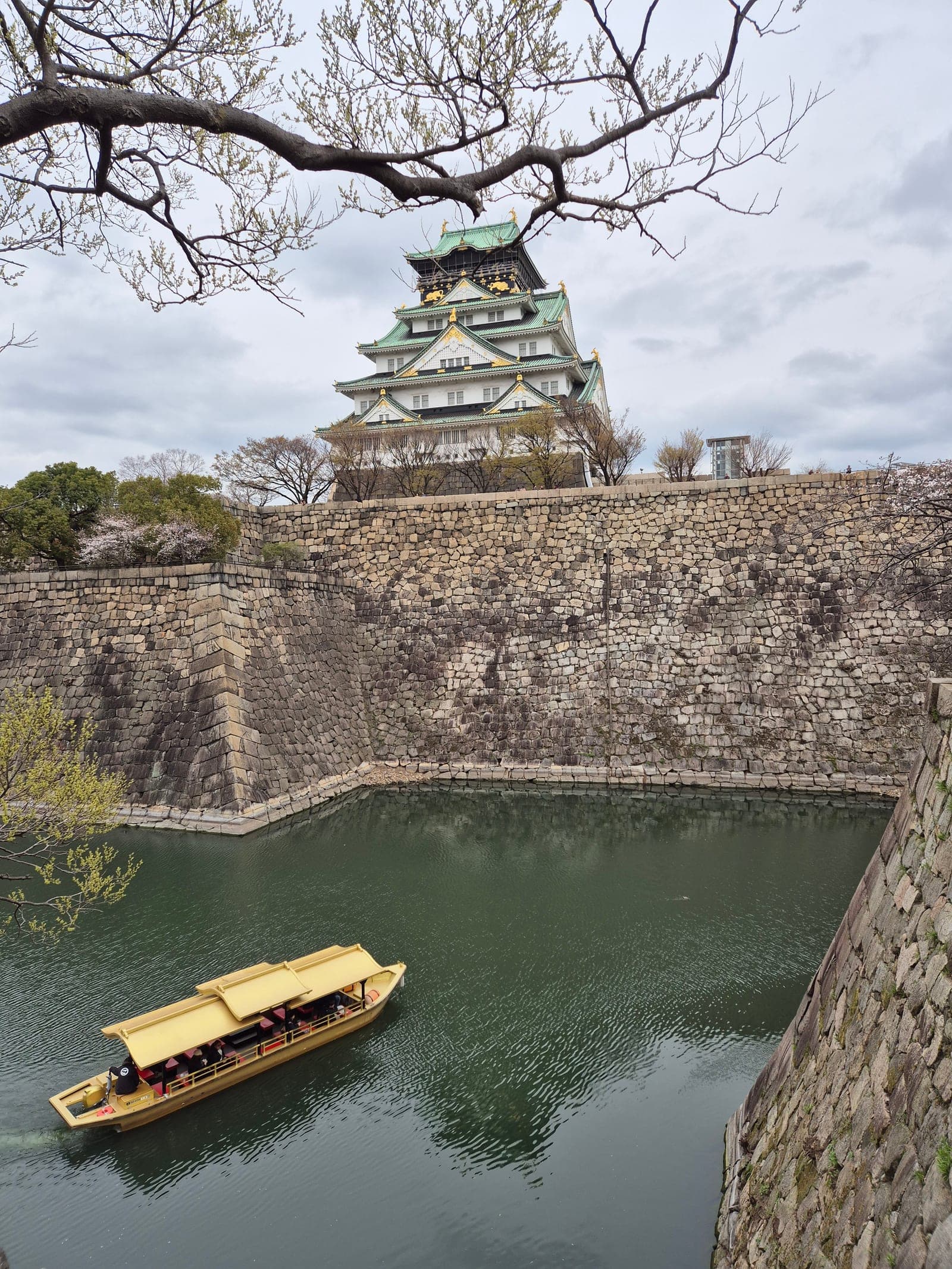 La torre principal del Castillo de Osaka sobre sus murallas de piedra, con un barco turístico dorado navegando por el foso